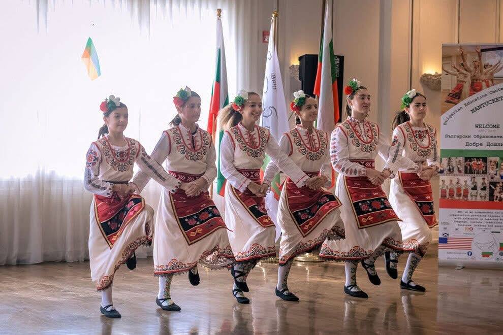 Six dancers in embroidered folk costume mid-step in front of Bulgarian and Ukrainian flags