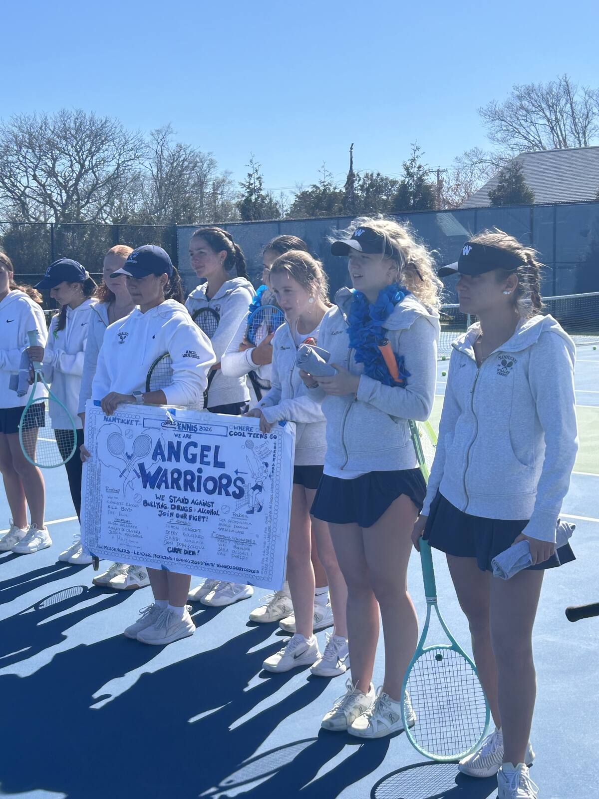 The girls' tennis team gathered on court around a hand-painted Angel Warriors sign